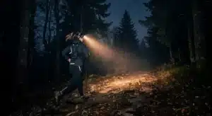 Photo réaliste d'un traileur vu de dos ou de profil, courant de nuit sur un sentier forestier de moyenne montagne (ambiance Vosges), faisceau lumineux net et visible projeté par sa lampe frontale sur le chemin devant lui